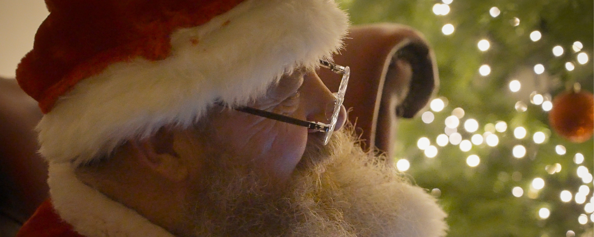 Santa Claus in his red hat and glasses beside a lit Christmas tree